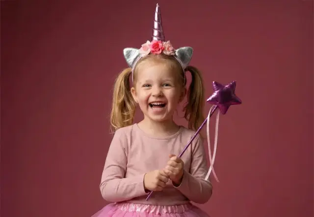 Dziecko ma na sobie opaskę z jednorożcem i różową tiulową spódniczkę oraz trzyma fioletową różdżkę z gwiazdką na tle delikatnego różu.